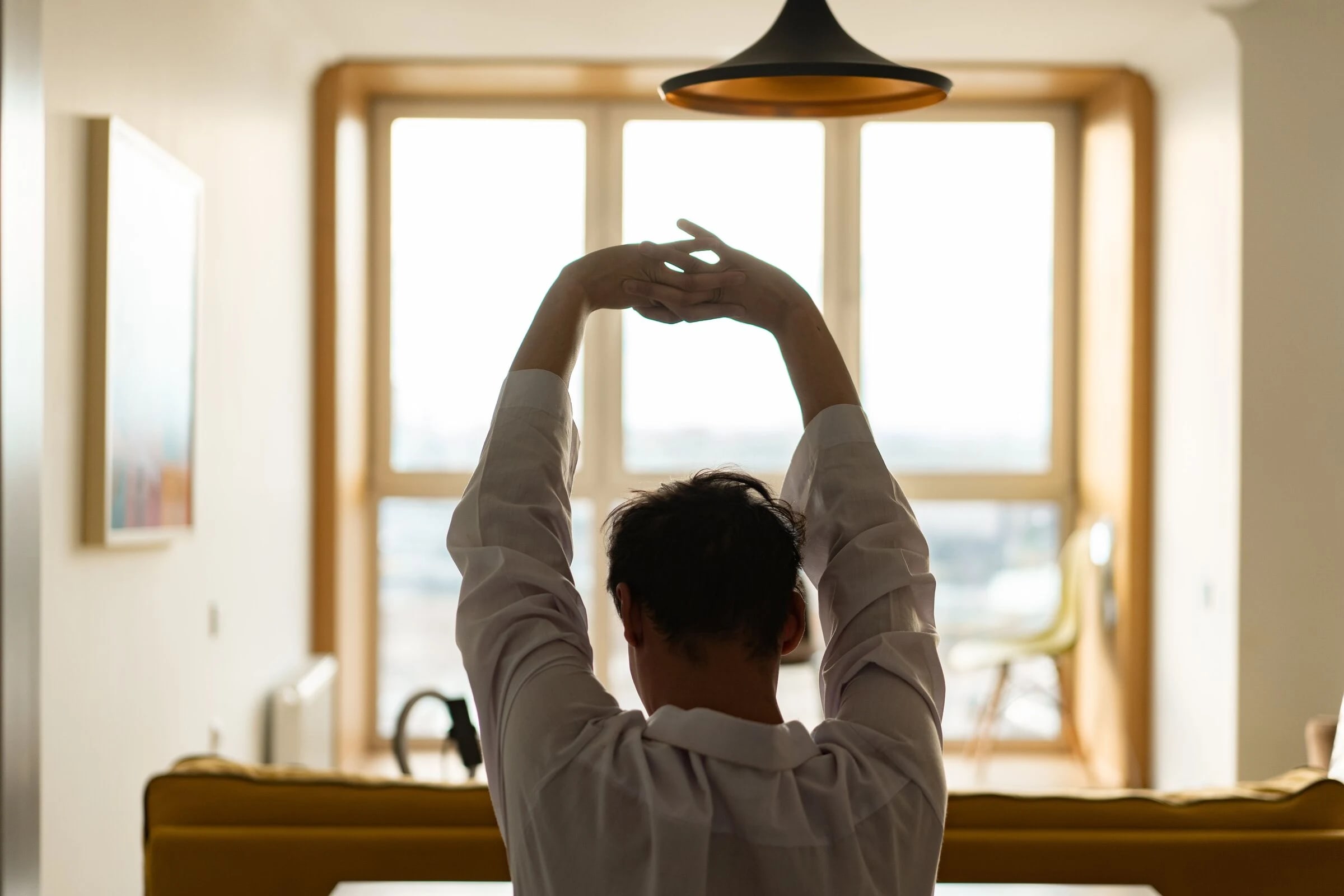 A person stretching at a bright window in the morning, seen from behind